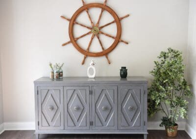 Decorative nautical-themed wall display with a ship’s wheel above a gray cabinet adorned with small vases and plants at Charter Senior Living of Paris, TN.