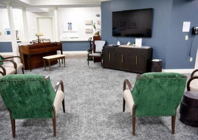 Spacious lounge area with green upholstered chairs, a piano, large TV, and dark wood cabinet at Charter Senior Living.