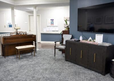 Common area at Charter Senior Living of Paris, TN, featuring a wooden piano with a cushioned bench, a flat-screen TV mounted on a navy accent wall, and a dark wood cabinet with patriotic décor.