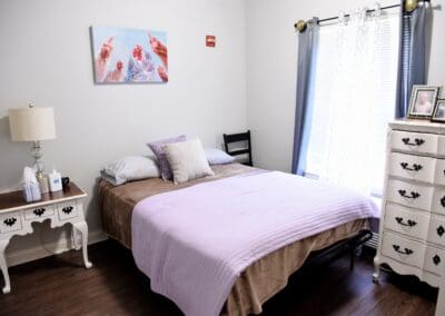 Cozy senior living bedroom with a lavender bedspread, white vintage furniture, and natural light at Charter Senior Living of Paris, TN