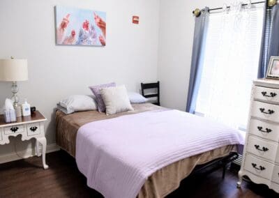 Cozy senior living bedroom with a lavender bedspread, white vintage furniture, and natural light at Charter Senior Living of Paris, TN