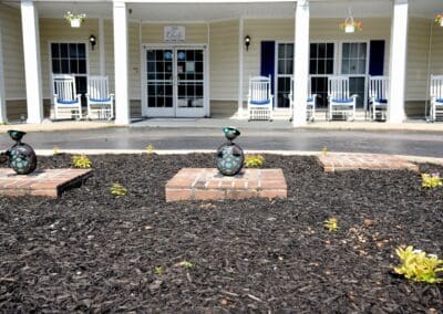 Front entrance of Charter Senior Living of Paris in Paris, TN, featuring a landscaped garden with decorative ornaments and white rocking chairs on the porch